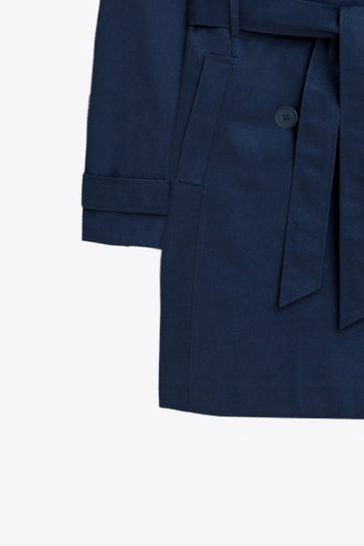 Abrigo trench corto de fit recto, con cuello solapa, bolsillos delanteros con vivos, detalle de bata trasera y manga larga. Trabillas, cinturón amarrable del mismo tejido y cierre frontal con doble cruce de botones en contraste. Detalle de trabilla y botón en hombros, bata trasera, cuello y puños.