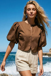 Blusa corta de fit recto, cuello redondo, manga corta amplia globo con volumen en borde, cierre frontal con broches ocultos y bajo curveado frontal.