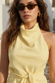 Blusa de fit recto, halter con cuello fluido, sin mangas, lazo amarrable en cintura, cierre posterior con botón y ojal.