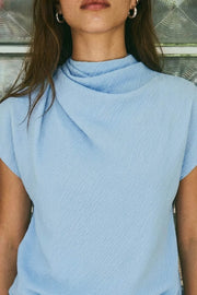 Blusa de cuello alto fluido, plisados en hombros, manga corta seguida y fit recto. Cierre posterior con botón y ojal.