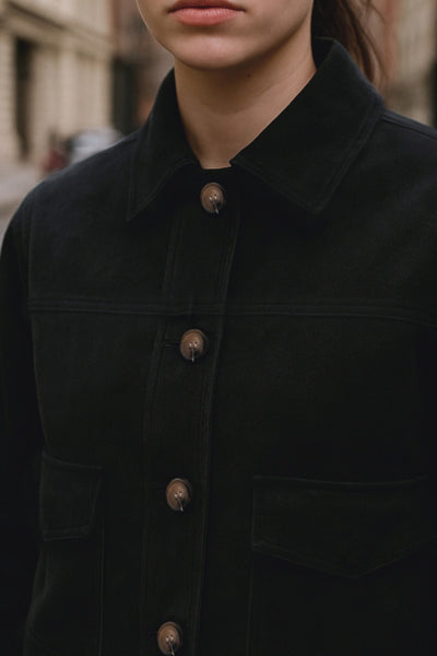 Chamarra corta fluida de gamuza, manga larga con puño abotonado, cuello camisero, bolsillos frontales de parche y cierre frontal con hilera de botones en contraste.