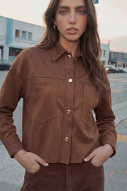 Chamarra corta de gamuza, fit oversized, cuello camisero, manga larga con puño abotonado, corte de bata frontal y posterior, bolsillos frontales de parche y cierre frontal con hilera de botones en contraste.