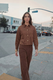 Chamarra corta de gamuza, fit oversized, cuello camisero, manga larga con puño abotonado, corte de bata frontal y posterior, bolsillos frontales de parche y cierre frontal con hilera de botones en contraste.