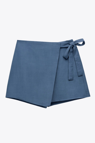 Falda short corto de cintura alta, corte en a, sobrefalda con cruce frontal, lazo lateral amarrable y cierre posterior con cremallera oculta.