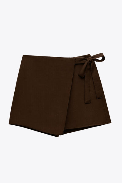 Falda short corto de cintura alta, corte en a, sobrefalda con cruce frontal, lazo lateral amarrable y cierre posterior con cremallera oculta.