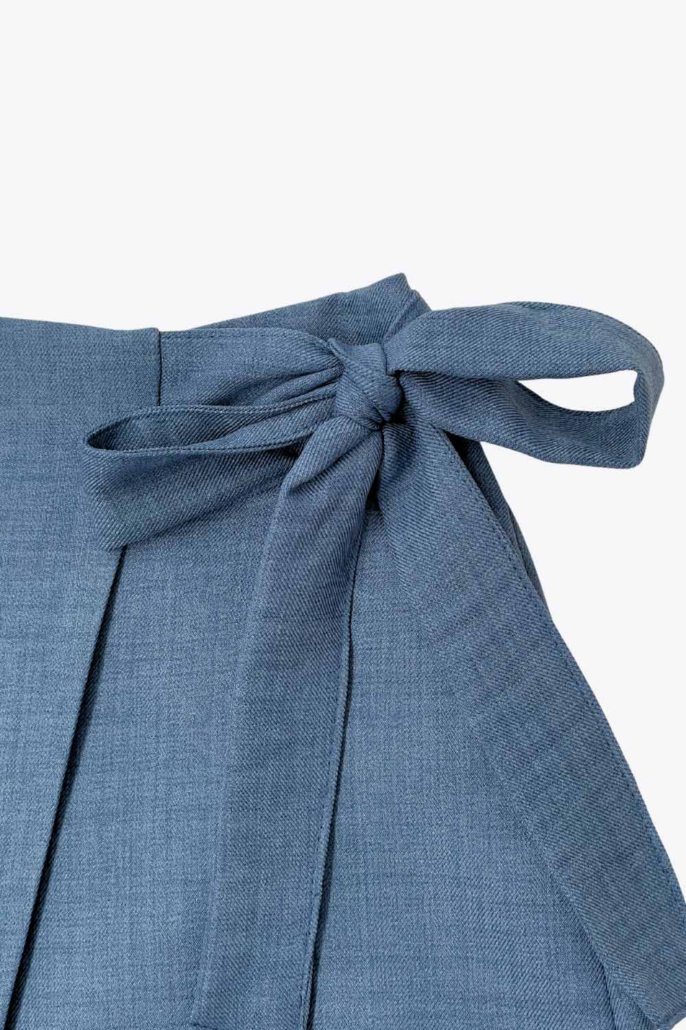 Falda short corto de cintura alta, corte en a, sobrefalda con cruce frontal, lazo lateral amarrable y cierre posterior con cremallera oculta.