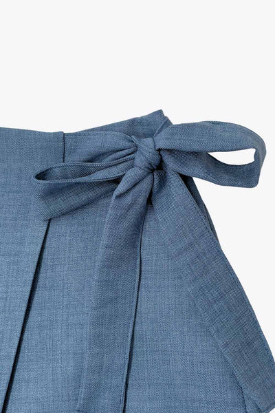 Falda short corto de cintura alta, corte en a, sobrefalda con cruce frontal, lazo lateral amarrable y cierre posterior con cremallera oculta.