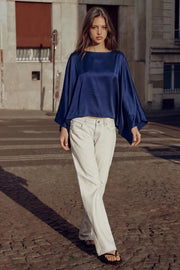 Blusa satinada tipo capa de fit fluido, manga larga amplia, cuello barco, bajo asimétrico y textura en tejido.