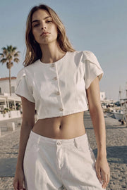 Blusa corta de fit recto, manga corta amplia con abertura, olanes y plisados en hombros, cortes frontales con pinzas, cuello redondo y cierre frontal con hilera de botones en contraste.