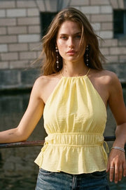 Top corto de cuello halter con plisados, tirantes delgados, detalle de rib con plisados y lazos laterales en costados, peplum con olanes en bajo y cierre posterior con botón y ojal.
