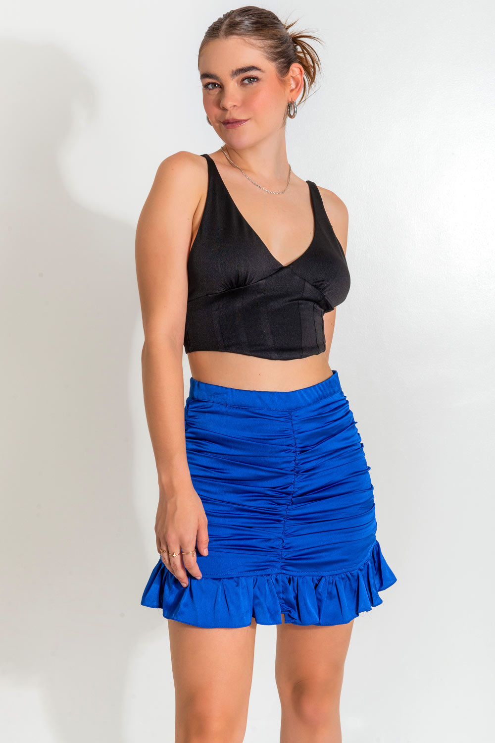 Falda corta satinada de fit ajustado, cintura alta con pretina elástica, fruncido frontal central y lateral. Bajo con olanes en borde y cierre lateral con cremallera oculta.