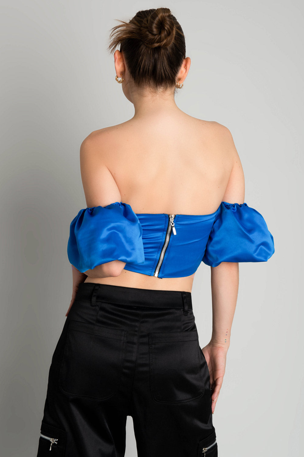 Top corset satinado de escote con detalles plisados, costuras frontales, bajo v frontal, hombros descubiertos y manga corta abullonada con olanes. Cierre con cremallera visible en contraste. 